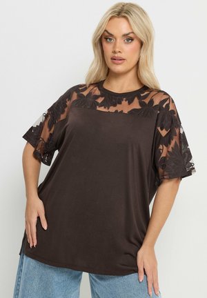 Kvinde iført en løs, mørkebrun top med gennemsigtige blomstrede blonder på korte ærmer og øvre bryst, kombineret med lyseblå jeans, stående med hånden på hoften.