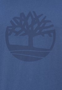 Μπλε βαμβακερό T-shirt με σκούρο μπλε κυκλικό λογότυπο δέντρου, με γεωμετρικά κλαδιά που επεκτείνονται από το κέντρο, πάνω σε υφασμένο φόντο.