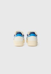 Baskets avec une tige crème, accents bleus et une semelle en caoutchouc, présentant une texture lisse et un design minimaliste, étiquetées "TOMMY JEAN".