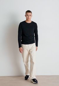 Melna garā piedurkne krekls, krēmkrāsas bikses un melnas kedas. Modelis stāv neitrālā telpā, demonstrējot minimālu apģērbu.