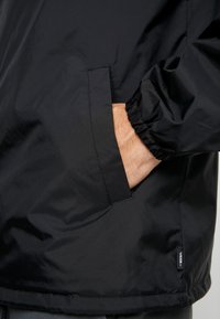 Giacca a vento nera con polsini elasticizzati, una tasca laterale e un'etichetta tessuta sull'orlo. Tessuto morbido, materiale leggero.