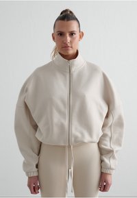 Beige mikina na zip s oversized střih, vysokým límcem a žebrovanými manžetami, v kombinaci s těsnými béžovými legínami. Měkká, texturovaná látka.
