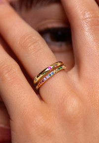 Anelli d'oro impilati su un dito, con gemme colorate in rosa, blu e verde, caratterizzati da superfici lisce e lucide.
