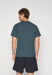 Mørk teal t-skjorte med korte ermer, rund hals og avslappet passform, sammen med sorte shorts, vist bakfra.