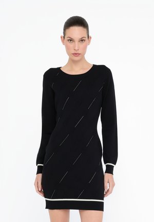 Donna che indossa un vestito lungo a maniche nere aderente con sottili motivi diagonali e bordi bianchi su polsini e orlo, in piedi contro uno sfondo bianco.
