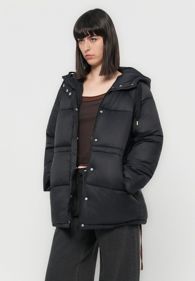 Schwarze Puffjacke mit Kapuze, gepolstertem Design, vorderen Druckknopfverschlüssen und Zugbandakzenten. Trägt man über einem braunen Oberteil und grauen Hosen.