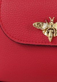 Rode handtas van getextureerd leer met een afgeronde klep en gouden bijvormige hardware-accent. Heeft stiksels langs de rand.