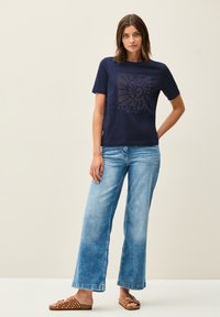 T-shirt en coton bleu marine à manches courtes, dotée d'un design de logo géométrique en couleurs claires. Associée à un jean large bleu clair et des sandales marron.