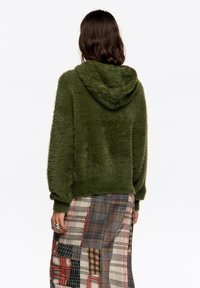 Sudadera verde peluda con capucha, mangas largas y un corte holgado, combinada con una falda de cuadros estampada.