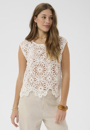 Junge Frau, die ein weißes, floral gehäkeltes ärmelloses Top und beige Hosen trägt, accessoriert mit goldenen und bernsteinfarbenen Armbändern und Ohrringen.