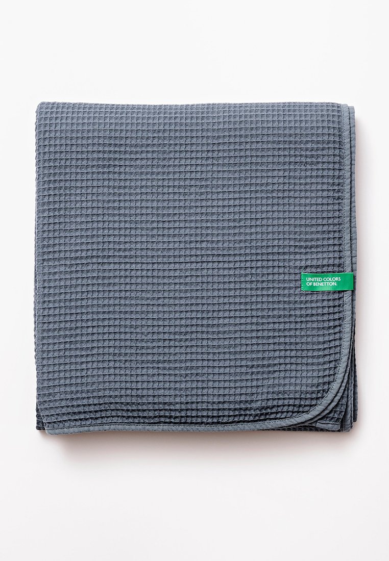 Manta gris de textura con un patrón de gofre, que presenta una etiqueta verde con la inscripción "UNITED COLORS OF BENETTON." Doblada ordenadamente.