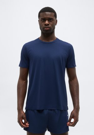 Muž stojaci čelom vpred, oblečený v jednoduchom tmavomodrom tričku s krátkym rukávom a zodpovedajúcich šortkách na bielom pozadí.