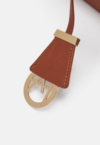 Sangle de sac en cuir marron avec un accent métallique doré, présentant un détail de logo circulaire et des bords cousus. Finition texturée observée.