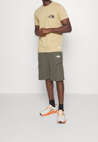 Muž stojící v béžovém tričku The North Face, olivově zelených šortkách s logy, černých ponožkách a bílých teniskách s oranžovými podrážkami a tkaničkami.