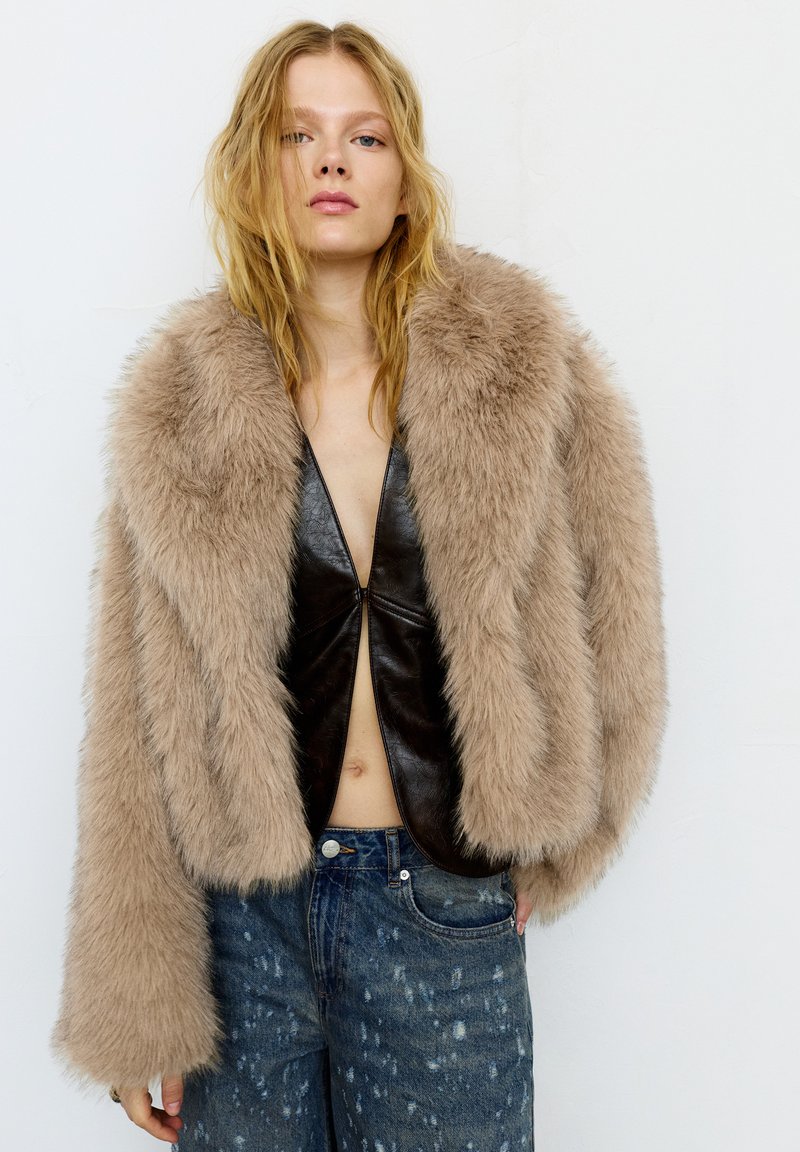 Jeune personne aux longs cheveux blonds portant une veste en fausse fourrure marron clair par-dessus un haut en cuir noir et un jean bleu déchiré, debout contre un mur blanc.