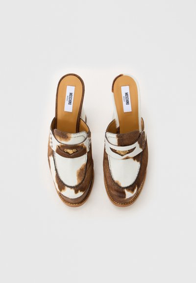Mule slip-on cu imprimeu de vacă maro și alb, cu branțuri bej și embellishmenturi aurii în formă de inimă, etichetate Moschino Milano.