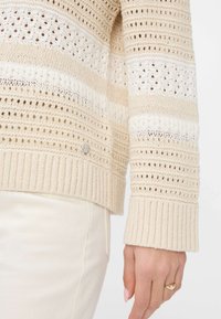 Maglione beige lavorato a maglia con strisce a giorno, polsini a costine e un piccolo dettaglio metallico di lato. Texture morbida e design leggero.