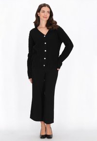 Zwarte gebreide cardigan met een V-hals, voorzien van vijf grote zilveren knoppen, gepresenteerd over zwarte wijde broek en zwarte hakken.