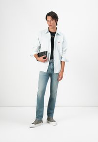 Ungdoms mann i lys skjorte, svart T-skjorte, blå jeans, grå sneakers, som holder en svart veske mot en ensfarget hvit bakgrunn.