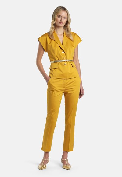 Femme portant un blazer jaune moutarde à manches courtes avec boutons, un pantalon slim assorti, une ceinture dorée et des escarpins dorés à bout pointu, debout la main dans la poche.