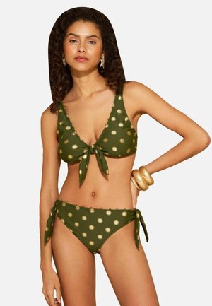 Femeie cu păr creț purtând un bikini verde cu modele aurii de soare, cercei în formă de stea și brățări mari aurii la încheietura unei mâini.