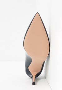 Chaussure stiletto noire à talon haut vue de la semelle avec un bout pointu et une semelle beige lisse sur fond blanc.