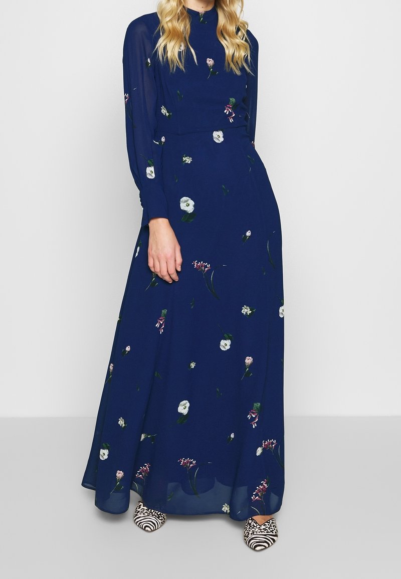 Robe maxi à manches longues imprimée de motifs floraux sur tissu bleu marine. Comprend un col montant, une jupe fluide et des manches transparentes. Associée à des chaussures à rayures noires et blanches.