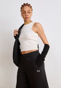 Bela rebrasta majica na bretele uparena s crnim trenirkom s malim Puma logotipom. Model drži crnu puffer jaknu, vidljive duge crne rukave.