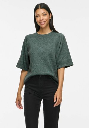 Jeune femme aux longs cheveux foncés portant un pull vert ample à manches courtes et un pantalon noir taille haute, souriante à la caméra.
