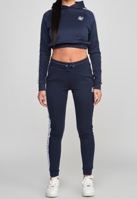 Žena oblečená v námořnickém modrém zkráceném svetru s kapucí a odpovídajících teplákách s bílým logem a pruhy, stojící proti šedému pozadí.