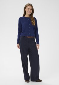 Marineblauer Cardigan mit Knöpfen, kombiniert mit marineblauen Hosen mit weitem Bein. Verfügt über eine glatte Textur und zwei Fronttaschen. Akzent durch braunen Gürtel.