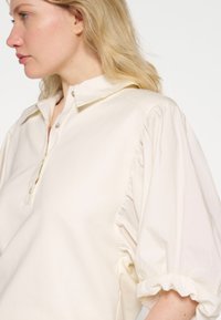 Crème kleurige shirt met een knoopsluiting, een klassieke kraag en pofmouwen met gerimpelde manchetten. Gemaakt van lichtgewicht stof.