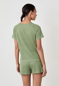Woman with braided hair wearing light green short-sleeve shirt and matching shorts, standing sideways against plain light background.