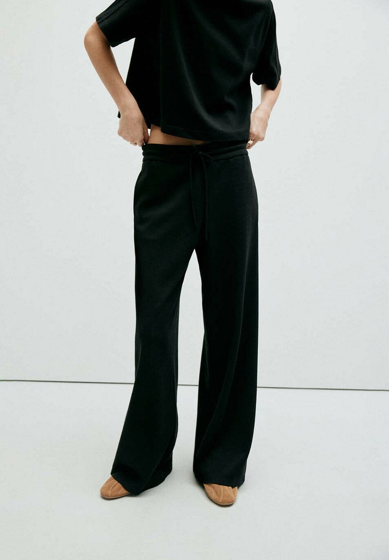 Personne portant un pantalon large noir, un haut noir à manches courtes et des chaussures marron clair, ajustant le pantalon à la taille sur un fond uni.