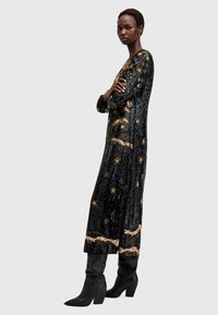 Rochie neagră din paiete cu stele și modele brodate în aur, cu mâneci lungi, o siluetă dreaptă și o tăietură laterală, asortată cu cizme negre până la genunchi.