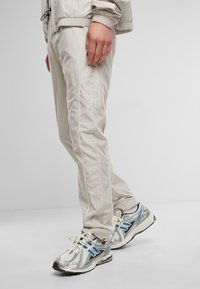 Lichtbeige sportbroek met zijpiping, gecombineerd met zilveren en blauwe sneakers. De stof lijkt glad met een aangesloten ontwerp.