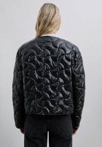 Chaqueta acolchada negra con un acabado brillante, que presenta un patrón de olas, cuello acanalado y un ajuste relajado, combinada con jeans oscuros.