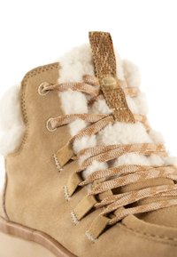 Bota de ante beige hasta el tobillo con forro de piel suave, cordones marrones trenzados y una pestaña texturizada. Cuenta con una suela de goma y detalles de costura visibles.