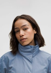 Vaaleansininen takki, jossa on korkea kaulus ja nappi-detail. Pehmeä, teksturoitu materiaali, rento istuvuus ja näkyvät saumat.