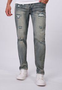 Hellblaue, abgenutzte Jeans mit Rissen und ausgefransten Kanten, im Slim-Fit-Design und kontrastierenden Nähten, kombiniert mit weißen Sneakers.