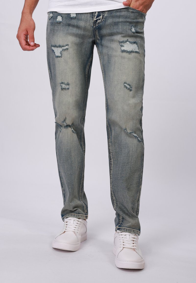 Hellblaue, abgenutzte Jeans mit Rissen und ausgefransten Kanten, im Slim-Fit-Design und kontrastierenden Nähten, kombiniert mit weißen Sneakers.