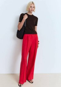 Chemise en tricot marron associée à un pantalon large rouge en lin taille haute. Portant un sac fourre-tout tissé noir, se tenant contre un fond clair.