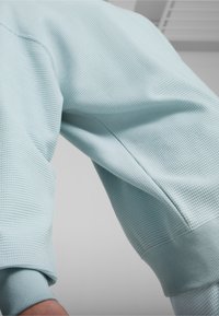 Ljusblå texturerad långärmad tröja med ribbade muddar, bärs på armen mot en neutral bakgrund.