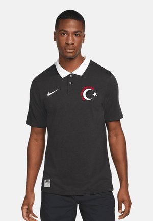 Mann trägt ein schwarzes Nike-Poloshirt mit weißem Kragen und einem Halbmond- und Stern-Logo auf der linken Brust, steht vor einem schlichten Hintergrund.