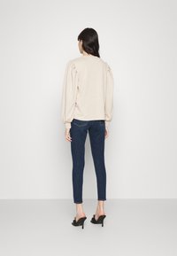 Beige tröja med puffärmar, rundad halsringning, kombinerad med mörkblå skinny jeans och svarta högklackade mules, sedd bakifrån.