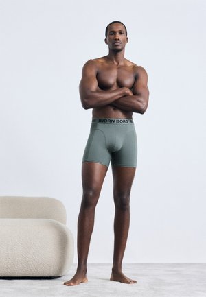 Green boxer briefs with black waistband, featuring the "BJÖRN BORG" logo. Model stands with arms crossed, showcasing a fitted design and smooth texture.
