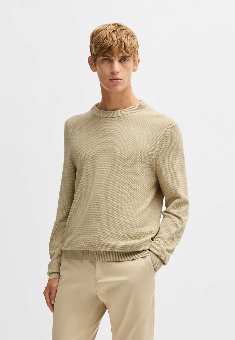 Beige neulepaita, jossa on pyöreä kaula-aukko ja pitkät hihat, ja joka sisältää teksturoitua kangasta sekä ribbaavia hihansuita ja helmaa. Yhdistettynä beigeihin housuihin.