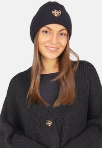 Un bonnet en tricot noir avec un patch en forme de cœur léopard. Le mannequin porte un cardigan noir à finition texturée et un bouton rond en métal.