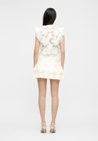 Mujer con cabello largo y oscuro, vestida con un top sin mangas de encaje blanco y una falda corta a juego, de pie de espaldas contra un fondo liso.