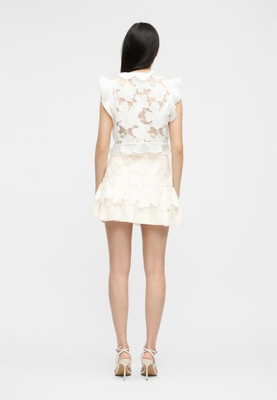 Γυναίκα με μακριά σκούρα μαλλιά, που φορά λευκό δαντελωτό αμάνικο τοπ και ταιριαστή κοντή φούστα, όρθια με την πλάτη γυρισμένη σε ένα απλό φόντο.